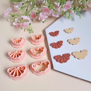 Peut inclure: Six moules en plastique rose pour la fabrication de boucles d'oreilles en argile en forme d'éventail. Deux boucles d'oreilles en argile finies sont présentées, l'une en marron et l'autre en beige.