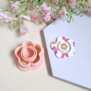 Peut inclure: Un emporte-pièce en forme de fleur rose clair à côté d'une pièce en pâte polymère en forme de fleur décorée. La pièce en argile a une base blanche avec des accents floraux roses et dorés. Des fleurs roses sont en arrière-plan.