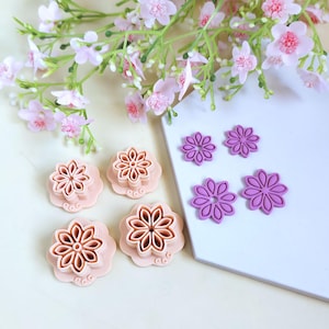 Peut inclure: Diverses découpoirs à biscuits en forme de fleurs, roses et violets, disposés sur une surface blanche. Les découpoirs présentent différentes tailles et motifs, certains avec des détails de pétales complexes. Des brins de fleurs roses en arrière-plan.
