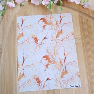 May include: A sheet of marbled paper with a white background and a swirling pattern of orange and gold. The paper has a subtle texture and is perfect for crafting or scrapbooking. The text "CamClayCo" is printed in the bottom right corner.