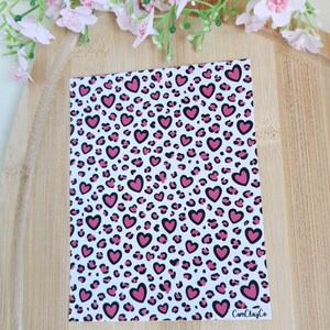 Peut inclure: Une feuille rectangulaire avec un fond blanc présentant un motif de cœurs roses soulignés en noir, entrecoupés de taches d'imprimé léopard. Le texte "CamClayCo" est visible dans le coin inférieur droit.
