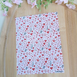 Könnte beinhalten: Eine rechteckige Karte mit weißem Hintergrund, bedeckt mit einem Muster aus roten und rosa Herzen und kleinen Blättern. Die Karte liegt auf einer Holzoberfläche, mit rosa Blüten im Hintergrund. Der Text "CamClayCo" ist sichtbar.