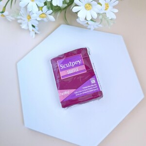 Puede incluir: Un paquete de arcilla polimérica Sculpey Souffle de color morado y blanco. El paquete está etiquetado con el nombre de la marca, el nombre del producto y el peso: 1,7 oz (48 g).