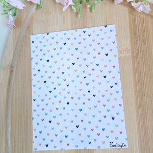 Könnte beinhalten: Eine weiße Karte mit einem sich wiederholenden Muster aus kleinen, bunten Herzen in Rot, Blau, Rosa, Gelb und Schwarz. Die Karte liegt auf einer Holzoberfläche, mit rosa Blumen im Hintergrund. Der Text "CamClayCo" ist sichtbar.