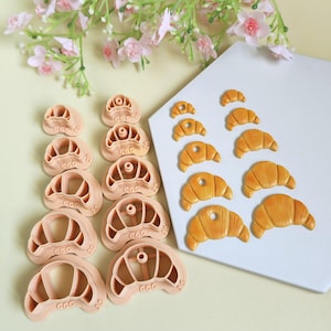 May include: A set of ten peach-colored plastic croissant-shaped clay cutters in varying sizes, paired with ten corresponding golden-brown croissant clay charms. The cutters feature the text CamClayCo on the base and size labels on the side.