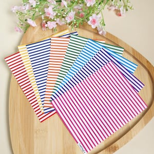 Op de afbeelding: Een verzameling kleurrijke kaarten met strepen, gerangschikt op een houten dienblad, met roze bloemen op de achtergrond. De kaarten hebben verticale strepen in verschillende kleuren, waaronder rood, geel, blauw, oranje, groen en paars.