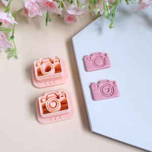 Peut inclure: Deux emportes-pièces en plastique rose en forme d'appareil photo et deux emportes-pièces en plastique rose en forme d'appareil photo avec un détail en forme de cœur. Les emportes-pièces sont sur une surface blanche.