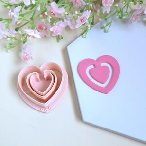 Könnte beinhalten: Ein Set aus drei herzförmigen Ausstechern in Rosa und ein einzelner herzförmiger Ausschnitt in Rosa. Die Ausstecher sind ineinander verschachtelt, mit einem kleineren Herzen in jedem größeren. Der Ausschnitt ist ein durchgehendes rosa Herz mit einer kleineren Herzform im Inneren.