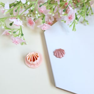 Puede incluir: Una herramienta de manualidades en forma de concha de color rosa claro y un adorno en forma de concha se encuentran sobre una superficie blanca. Flores y vegetación rosas en el fondo. La herramienta tiene una base circular con un diseño festoneado.