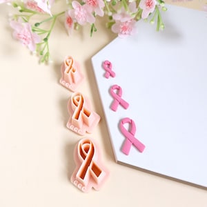 Puede incluir: Cuatro cortadores de galletas de cinta rosa de plástico de diferentes tamaños y tres cintas de fondant rosa de diferentes tamaños sobre una superficie blanca.