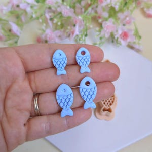 Può includere: Quattro piccoli pezzi artigianali a forma di pesce azzurro, con dettagli di squame e un foro in alto. È visibile anche uno stampo a forma di pesce marrone chiaro. Lo sfondo comprende fiori rosa e una superficie bianca.