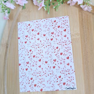 Peut inclure: Une carte rectangulaire avec un motif de coeurs rouges et de feuilles sur fond blanc. La carte est posée sur une surface en bois, avec des fleurs roses en arrière-plan. Le texte "CamClayCo" est visible.