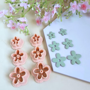 Könnte beinhalten: Eine Sammlung von rosa und grünen Ausstechern in Blumenform. Die rosa Ausstecher sind auf einer weißen Oberfläche angeordnet, mit ein paar grünen Blumenformen in der Nähe. Rosa Blumen und Grünzeug befinden sich im Hintergrund.