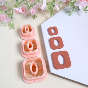 May include: Pink square-shaped polymer clay cutters and three brown square-shaped clay pieces with a hole in the center. The cutters are in different sizes. The image also includes pink flowers and a white surface.