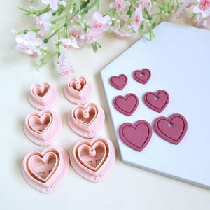 Könnte beinhalten: Eine Sammlung rosa herzförmiger Ausstecher und herzförmiger Tonstücke. Die Ausstecher sind ineinander verschachtelt, und die Tonherzen sind in verschiedenen Größen. Ein Zweig rosa Blüten im Hintergrund.