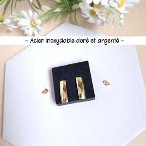 May include: Gold-colored rectangular earrings displayed on a black surface, with a white geometric background. The earrings are paired with gold-colored earring backs. The image also includes a sprig of white daisies.