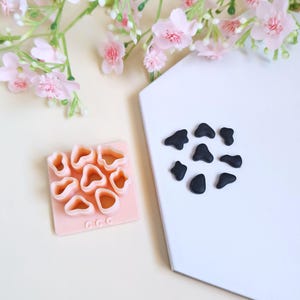 Può includere: Un taglierino in argilla polimerica rosa chiaro con più forme irregolari, accanto a una collezione di pezzi di argilla nera. Gli oggetti sono su una superficie bianca, con fiori rosa sullo sfondo. Il taglierino ha le lettere "CCC".