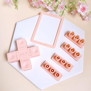 Puede incluir: Un conjunto de herramientas de artesanía de plástico rosa, que incluye una regla con medidas en milímetros, un cuadrado con un agujero en el centro y cuatro filas de pequeños círculos de diferentes tamaños. Las herramientas están etiquetadas con "CCC" y "mm".