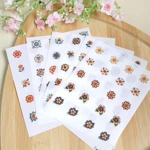 Peut inclure: Feuilles d'autocollants floraux avec divers motifs de fleurs colorées. Les autocollants présentent des fleurs orange, jaunes, bleues et violettes. Les autocollants sont sur du papier blanc, et le fond comprend une planche à découper en bois et des fleurs roses.