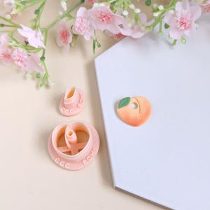 May include: Three pink plastic cookie cutters, one shaped like a heart, one shaped like a peach, and one shaped like a peach slice. The peach slice cutter has a green leaf detail. The cutters are on a white hexagonal tile.
