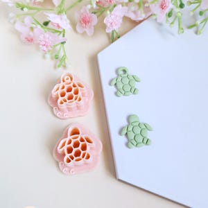 Peut inclure: L'image présente deux emporte-pièces roses en forme de tortue avec un motif en nid d'abeille, ainsi que deux petits charms verts en forme de tortue. Les objets sont disposés sur une surface blanche, avec des fleurs roses en arrière-plan. Le thème est l'artisanat ou la pâtisserie.