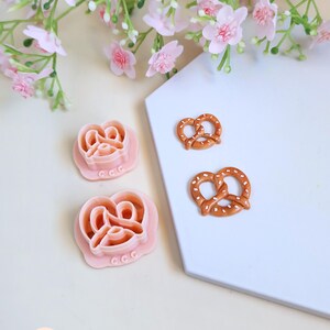 Peut inclure: Deux emporte-pièces roses en forme de bretzel et deux bretzels miniatures avec des éclats de sel blancs. Les emporte-pièces sont sur une surface blanche avec des fleurs roses en arrière-plan.
