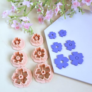 Peut inclure: Vue aérienne de découpoirs à biscuits en forme de fleurs roses et violettes. Ils sont disposés sur une surface blanche, avec une branche de fleurs roses en arrière-plan. Les découpoirs sont de différentes tailles et formes.