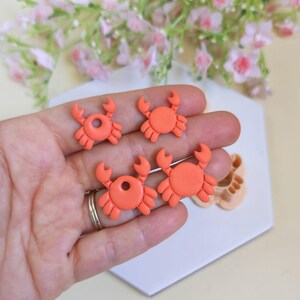 Puede incluir: Cuatro dijes de arcilla polimérica con forma de cangrejo naranja. Los dijes se sostienen en una mano, con una superficie blanca y gris al fondo. Flores rosas ligeramente borrosas al fondo. Los dijes miden aproximadamente 2,5 cm.