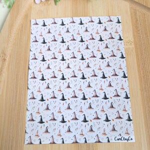 Può includere: Un foglio bianco con un motivo ripetuto di cappelli da strega neri e beige, con foglie autunnali. Il foglio è su una superficie di legno con fiori rosa sullo sfondo. Il testo "CamClayCo" è visibile.