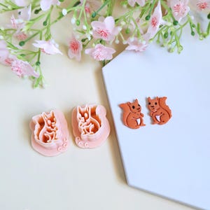 May include: Two orange squirrel-shaped cookie cutters and two squirrel-shaped cookies are displayed on a white surface. The cookie cutters are pink. Pink flowers and greenery are in the background. The cookie cutters are for making squirrel-shaped cookies.