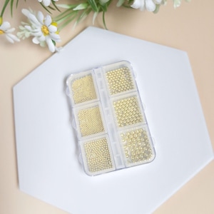 Puede incluir: Un recipiente de plástico transparente con seis compartimentos llenos de cuentas doradas de diferentes tamaños. El recipiente está sobre una superficie hexagonal blanca.