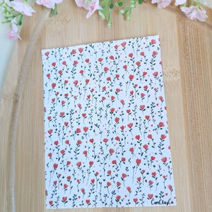 Könnte beinhalten: Eine rechteckige Karte mit einem floralen Muster aus roten Blumen und grünen Blättern auf weißem Hintergrund. Die Karte liegt auf einer Holzoberfläche, mit rosa Blüten im Hintergrund. Der Text "CamClayCo" ist sichtbar.