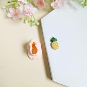 Peut inclure: Un moule en silicone rose en forme d'ananas, avec un petit charme ananas jaune et vert à côté.