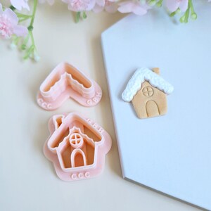 May include: Two pink plastic cookie cutters shaped like gingerbread houses. One cutter is closed and the other is open. The closed cutter has a white icing design on top. The cutters are on a light blue surface.