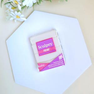 Puede incluir: Un paquete de arcilla polimérica Sculpey Premo, 57 g, con una etiqueta rosa y blanca. La arcilla está sobre una superficie hexagonal blanca, con flores blancas en el fondo. El paquete dice "Premium Oven-Bake Clay".