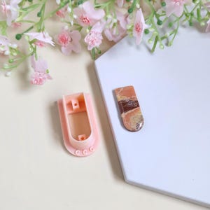 May include: A light pink cookie cutter and a rectangular stone with brown and tan patterns are displayed on a white surface. Pink flowers and greenery are in the background. The cookie cutter is in the shape of a rectangle with rounded edges.