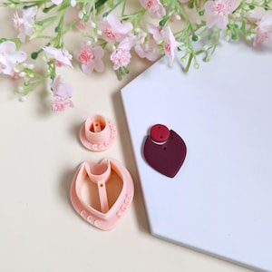Peut inclure: Un ensemble de découpeurs en pâte polymère rose clair avec un motif en forme de cœur, ainsi qu'un composant de boucle d'oreille en argile bordeaux avec un accent rouge. L'ensemble comprend un découpeur rond et un découpeur en forme de cœur, avec un arrangement floral en arrière-plan.
