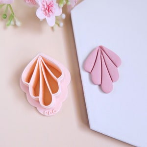 Könnte beinhalten: Ein rosa, plastikförmiger Keksausstecher in Form einer Muschel mit einem passenden rosa Tonabdruck. Der Ausstecher hat einen gewellten Rand und ein detailliertes Design.