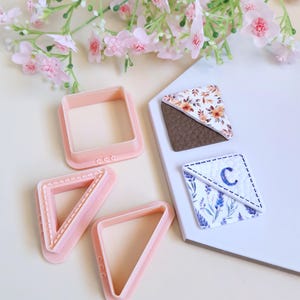 May include: A selection of pink geometric cookie cutters in square and triangle shapes, with two decorative clay pieces. One clay piece has a floral pattern, and the other features a lavender design with the letter "C". Pink flowers are in the background.