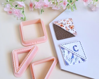 May include: A collection of pink geometric cookie cutters in square and triangle shapes, alongside two decorative clay pieces. One clay piece features a floral pattern, and the other has a lavender design with the letter "C". Pink flowers are in the background.