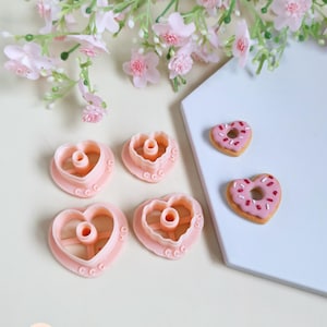 May include: A selection of pink heart-shaped cookie cutters and two heart-shaped donuts with pink icing and sprinkles. The cutters have a central hole and decorative details. Pink flowers are in the background.
