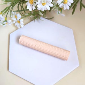 May include: A light-colored, wooden dowel rod rests on a white, hexagonal tile. White daisy flowers with yellow centers and green leaves are in the background. The dowel is a light tan color and has a ridged texture.