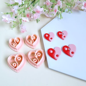 Peut inclure: Ensemble de découpoirs à biscuits roses en forme de cœur et de boucles d'oreilles rouges et roses en forme de cœur. Les découpoirs ont un motif yin-yang. Les boucles d'oreilles ont un motif yin-yang rouge et rose. Des fleurs roses sont en arrière-plan.