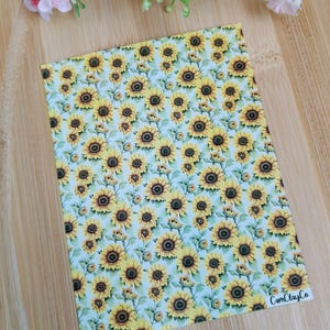 Può includere: Un foglio di pelle sintetica bianca con un motivo ripetitivo di girasoli gialli. Il testo "CamClayCo" è stampato nell'angolo in basso a destra.