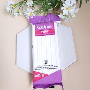Peut inclure: Un paquet d'argile polymère blanche Sculpey Premo, 227 g, avec un emballage violet et rose. Le paquet est posé sur une forme géométrique blanche, avec des fleurs blanches en arrière-plan.