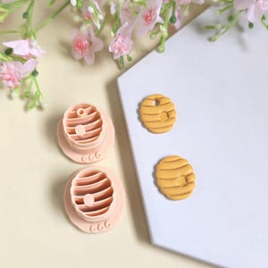 Peut inclure: Ensemble de découpoirs et d'embellissements en pâte polymère. Les découpoirs sont rose clair et en forme de ruche. Deux embellissements en forme de ruche jaunes sont également présents. Des fleurs roses sont en arrière-plan.