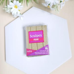 Könnte beinhalten: Eine 57 Gramm Packung goldfarbener, glitzernder Sculpey Premo Backofen-Modelliermasse.
