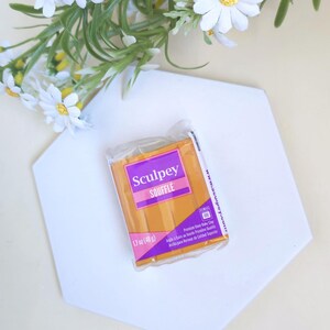 May include: A package of Sculpey Souffl&eacute; polymer clay, 1.7 oz (48 g), in a warm orange color, rests on a white hexagonal surface. The package has a purple label with the brand name. White daisy flowers with yellow centers and green foliage are in the background.