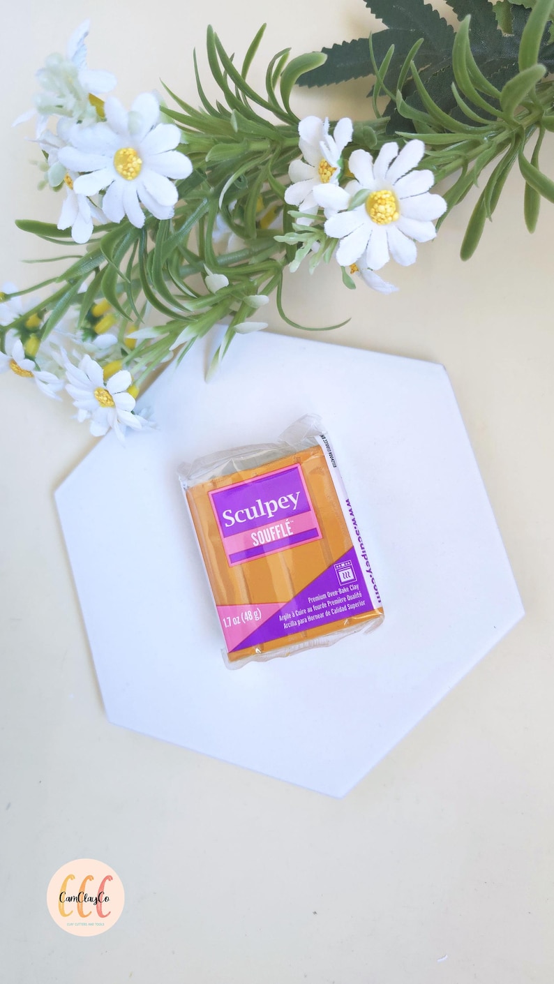 May include: A package of Sculpey Souffl&eacute; polymer clay, 1.7 oz (48 g), in a warm orange color, rests on a white hexagonal surface. The package has a purple label with the brand name. White daisy flowers with yellow centers and green foliage are in the background.