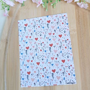 Peut inclure: Une carte rectangulaire avec un fond blanc orné d'un motif de cœurs rouges, roses et bleus, ainsi que de petits motifs floraux et de feuilles. Le texte "CamClayCo" est imprimé en bas à droite.